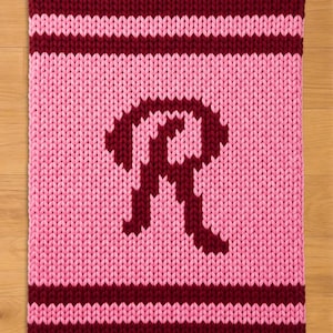 May include: A knitted blanket in shades of pink and burgundy. The blanket features a large letter "R" in burgundy on a pink background. Burgundy stripes border the top and bottom edges. The blanket is made of a thick, chunky knit.
