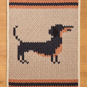 May include: A knitted rectangular rug featuring a dachshund dog design. The dog is black and peach, set against a beige background. The rug has a cream border and stripes in black and orange.