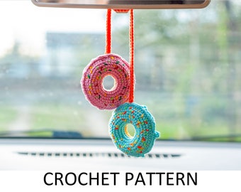 May include: Two crocheted donut shaped ornaments hanging from a rear view mirror in a car. The donuts are pink and blue with colorful sprinkles.
