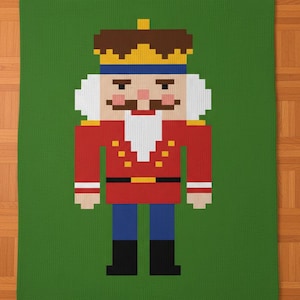 Puede incluir: Una manta verde con una imagen pixelada de un cascanueces. El cascanueces tiene una chaqueta roja con botones dorados, pantalones azules y botas negras. La corona es marrón con una parte superior dorada, y la cara tiene barba y bigote blancos.