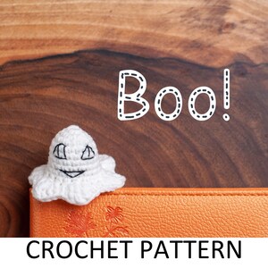 May include: A white crocheted ghost with a smiling face, sitting on an orange leather journal with the text "CROCHET PATTERN" and the word "Boo!" written in white with a dashed outline above the ghost.