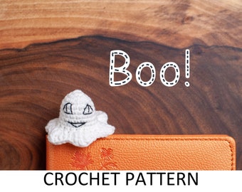 May include: A white crocheted ghost with a smiling face, sitting on an orange leather journal with the text "CROCHET PATTERN" and the word "Boo!" written in white with a dashed outline above the ghost.