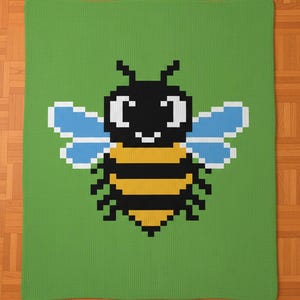 May include: A green blanket featuring a pixelated bee design. The bee has a black head and body stripes in yellow and black. The wings are light blue. The blanket is rectangular and rests on a wooden floor.