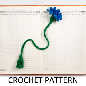 May include: A crochet bookmark with a blue flower and green stem and tassel. The bookmark is laying open on a page of a planner.