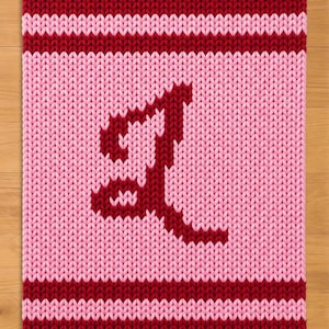 Peut inclure: Un article rectangulaire tricoté avec un fond rose et une grande lettre stylisée "J" de couleur rouge foncé. Les bords supérieur et inférieur sont rouge foncé uni, avec une bande rouge au milieu.