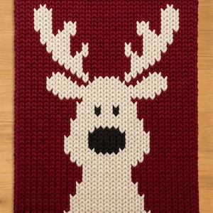 May include: A rectangular chunky knit blanket featuring a large, centered reindeer motif. The background is a deep burgundy red, while the reindeer is rendered in cream-colored yarn with a solid black nose and two small black eyes.