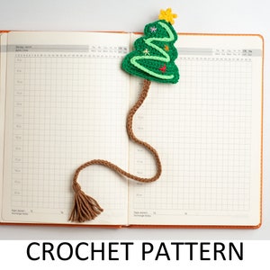 Könnte beinhalten: Ein grünes, gehäkeltes Weihnachtsbaum-Lesezeichen mit einem braunen Quasten. Das Lesezeichen befindet sich auf einem weißen karierten Notizbuch.