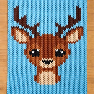 May include: A knitted blanket featuring a pixelated deer design. The deer has brown antlers, a brown body, and a beige face. The background is a solid light blue color. The blanket is made of a soft, chunky knit material.