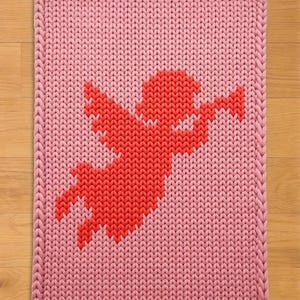 May include: A pink knitted rug with a red cupid design. The rug has a rectangular shape with a thick, braided border. The cupid is depicted in a pixelated style, with outstretched arms and wings, against a light pink background.