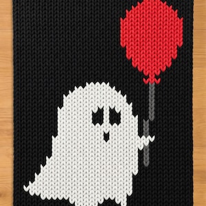 May include: A rectangular knit blanket featuring a black background with a white ghost character in the centre. The ghost has two black eyes and a small black mouth, and it is holding a red balloon attached to a grey string.