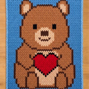 May include: A knitted square featuring a brown teddy bear holding a red heart. The bear has a light brown snout and ears, with black eyes. The background is a solid light blue, and the edges are also blue.