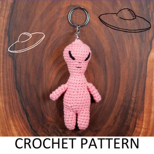 May include: Pink crocheted alien keychain with black eyes and a silver key ring. The alien is hanging against a wooden background with two hand-drawn UFOs.