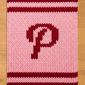 Pode incluir: Um artigo retangular de malha em tons de rosa e bordô. O artigo apresenta uma grande letra "P" bordô no centro. As bordas superior e inferior têm listras bordô e o fundo é rosa claro.