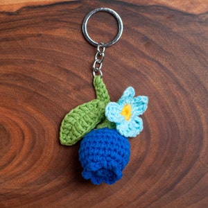 Op de afbeelding: Een gehaakte blauwe bloem met een groene steel en een lichtblauwe bloem met een geel hartje bevestigd aan een zilveren sleutelhanger.
