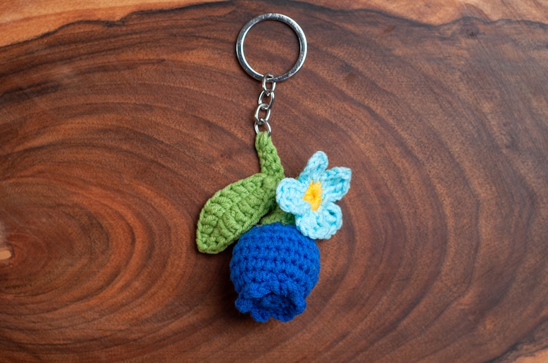Op de afbeelding: Een gehaakte blauwe bloem met een groene steel en een lichtblauwe bloem met een geel hartje bevestigd aan een zilveren sleutelhanger.