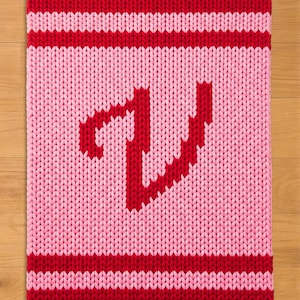 Puede incluir: Una manta de punto en tonos rojos y rosas. Los bordes superior e inferior son de color rojo liso, con una franja blanca entre ellos y el centro rosa. Una letra "J" roja pixelada está en el centro.