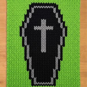 Puede incluir: Una manta de punto rectangular con un fondo verde lima brillante y un gran motivo de ataúd negro centrado. El ataúd está delineado en gris y contiene un símbolo de cruz gris en el centro, todo creado con una textura de punto gruesa y visible.