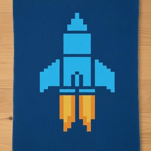 May include: A blue rectangular towel featuring a pixelated rocket ship design in shades of light blue and yellow. The rocket has a detailed pixelated design with flames at the bottom.