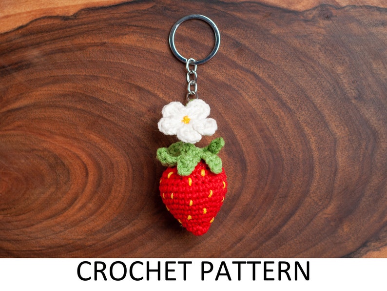 Puede incluir: Llavero de fresa tejido a crochet con una flor blanca y hojas verdes. El llavero est&aacute; unido a un llavero plateado.