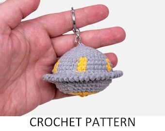 Puede incluir: Un llavero de OVNI tejido a crochet gris con detalles amarillos. El llavero se sostiene en una mano.