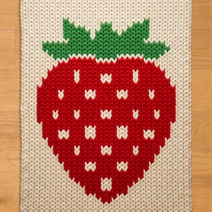 May include: A knitted square rug featuring a large red strawberry design with white seeds and green leaves. The strawberry is centered on a cream-colored background. The rug has a textured, woven appearance.