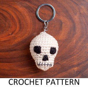 May include: A crocheted white skull keychain with black eyes. The keychain is attached to a silver key ring.