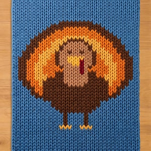 May include: A rectangular blue knit blanket featuring a central graphic of a turkey. The turkey is rendered in shades of brown, orange, and yellow, with a red wattle and yellow beak. The knit texture is consistent throughout the entire piece.