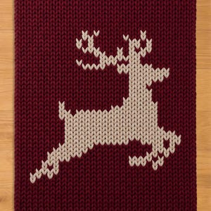 May include: A rectangular knitted blanket featuring a deep burgundy background with a large, centered cream-colored reindeer motif. The knit pattern is chunky and uniform, creating a textured surface that highlights the contrast between the dark red yarn and the light-colored deer silhouette.