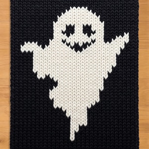 Könnte beinhalten: Ein rechteckiges Stricktextil mit einer großen, weißen Geisterfigur, die auf einem einfarbigen schwarzen Hintergrund zentriert ist. Der Geist hat zwei dreieckige Augen und einen gezackten, breiten Mund, alles in schwarzem Garn gearbeitet.