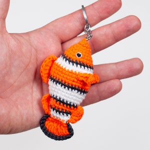 May include: A crocheted orange and white clownfish keychain with black stripes. The keychain is attached to a silver metal ring.