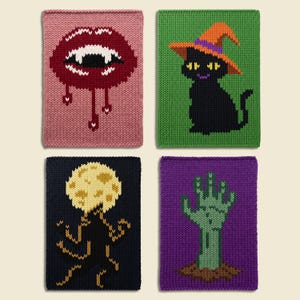 May include: Four rectangular knitted panels featuring distinct designs: a pink panel with a dark red vampire mouth, a green panel with a black cat wearing an orange witch hat, a black panel with a yellow moon, and a purple panel with a green hand.