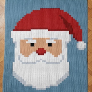 May include: A knitted blanket featuring a pixelated Santa Claus face. The design includes a red hat, white beard, and a light blue background. The blanket is a festive item for the holiday season.