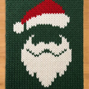 May include: A rectangular knitted blanket featuring a dark green background with a central graphic of a Santa Claus face. The design is created with white yarn for the beard and hat trim, and red yarn for the top of the Santa hat, all in a chunky knit.
