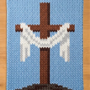 May include: A knitted blanket featuring a brown cross with white accents against a light blue background. The cross is set on a brown and gray base, with a light blue border. The blanket is made of a soft, textured material.