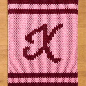 Peut inclure: Une couverture tricotée aux tons rose et bordeaux. La couverture présente un grand « A » bordeaux, centré sur un champ rose. Des rayures bordeaux bordent les bords supérieur et inférieur. La couverture est en grosse laine.