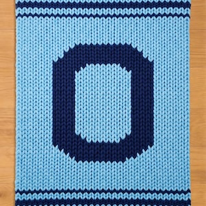 Puede incluir: Una alfombra de punto en tonos azules, con una gran letra "O" en azul marino sobre un fondo azul claro. La alfombra tiene rayas azul marino a lo largo de los bordes superior e inferior, creando un borde decorativo. La alfombra está hecha de hilo grueso y grueso.