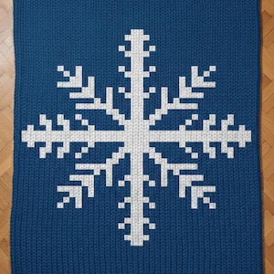 May include: A knitted blue blanket featuring a large white snowflake design. The snowflake is centered on the blanket and is made up of pixelated, geometric shapes. The blanket is rectangular and has rounded corners.