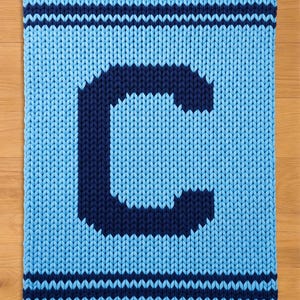 Op de afbeelding: Een gebreide deken in blauwtinten, met een grote marineblauwe letter "C" op een lichtblauwe achtergrond. Marineblauwe strepen bevinden zich aan de boven- en onderkant van de deken.