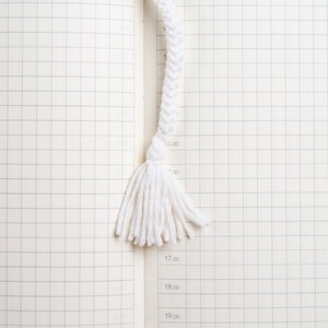 May include: A white braided rope bookmark with a tassel. The bookmark is on a white gridded notebook.