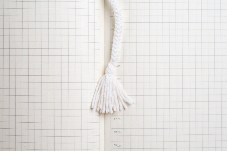 May include: A white braided rope bookmark with a tassel. The bookmark is on a white gridded notebook.