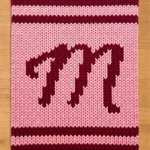 May include: A knitted rectangular rug in shades of pink and burgundy. The rug features a large burgundy letter "M" in the center, with burgundy stripes at the top and bottom. The background is a lighter pink.