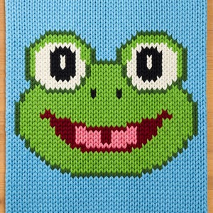 May include: A knitted square featuring a smiling frog face. The frog's face is green with white and black eyes, a pink mouth, and a dark red tongue. The background is light blue, and the edges are neatly finished.