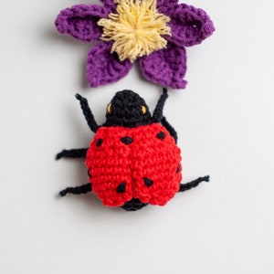 May include: A crocheted ladybug with black legs and red and black spots. The ladybug is sitting on a white background with a purple crocheted flower above it.