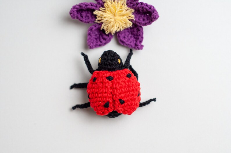 May include: A crocheted ladybug with black legs and red and black spots. The ladybug is sitting on a white background with a purple crocheted flower above it.