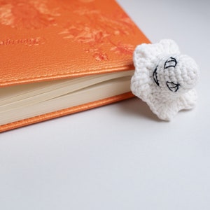 May include: A white crocheted ghost bookmark with black embroidered eyes and a smile, resting on an orange leather-bound journal.