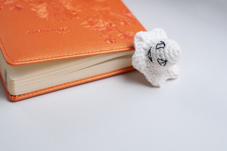 May include: A white crocheted ghost bookmark with black embroidered eyes and a smile, resting on an orange leather-bound journal.