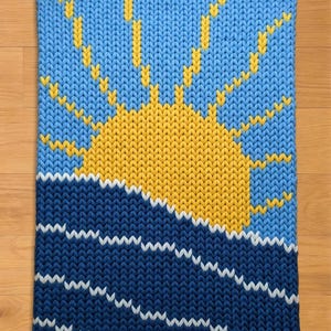May include: A knitted rectangular piece featuring a sun and sea design. The upper portion depicts a bright yellow sun with rays against a light blue sky. The lower section shows dark blue waves with white accents, creating a nautical theme.