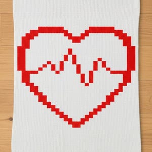 Può includere: Coperta bianca lavorata a maglia con un grande cuore rosso. Il cuore è pixelato, con una linea frastagliata al centro, che ricorda un battito cardiaco. La coperta è rettangolare e sembra essere fatta di materiale morbido.