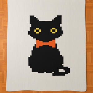 May include: A cream-colored blanket featuring a pixelated black cat with yellow eyes and an orange bow tie. The cat is centered on the blanket, which is displayed on a wooden floor.