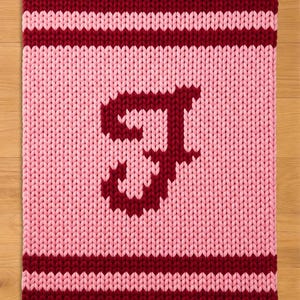 Könnte beinhalten: Eine gestrickte, rechteckige Decke in Rosa und Burgunderrot. Die oberen und unteren Ränder sind burgunderrot, mit einem burgunderroten Streifen in der Mitte. Ein großer burgunderroter Buchstabe "J" ist auf dem rosa Feld zentriert.